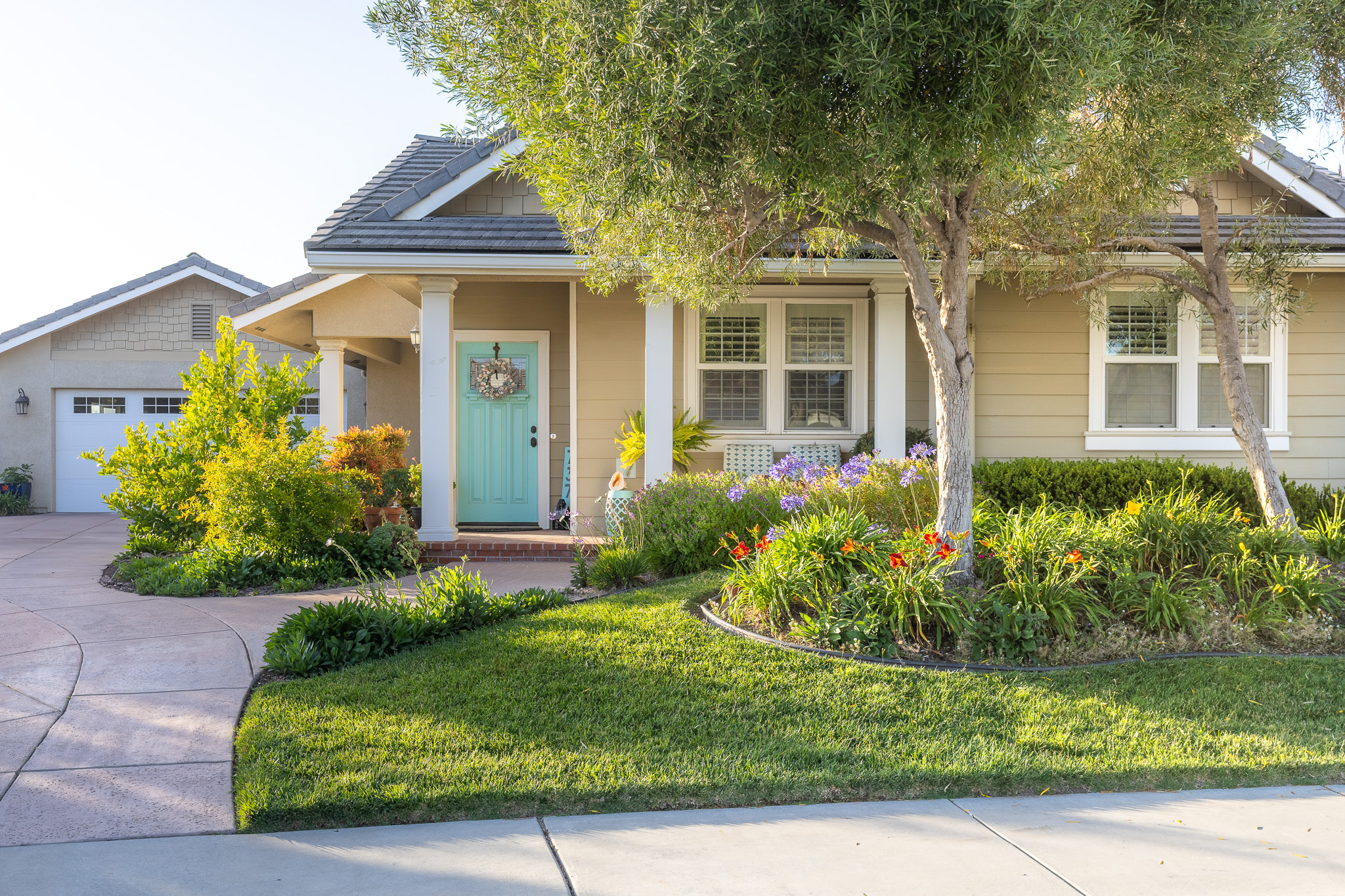 A craftsman-style home with a teal front door, seen across a green lawn and flowering garden in late-afternoon sun.